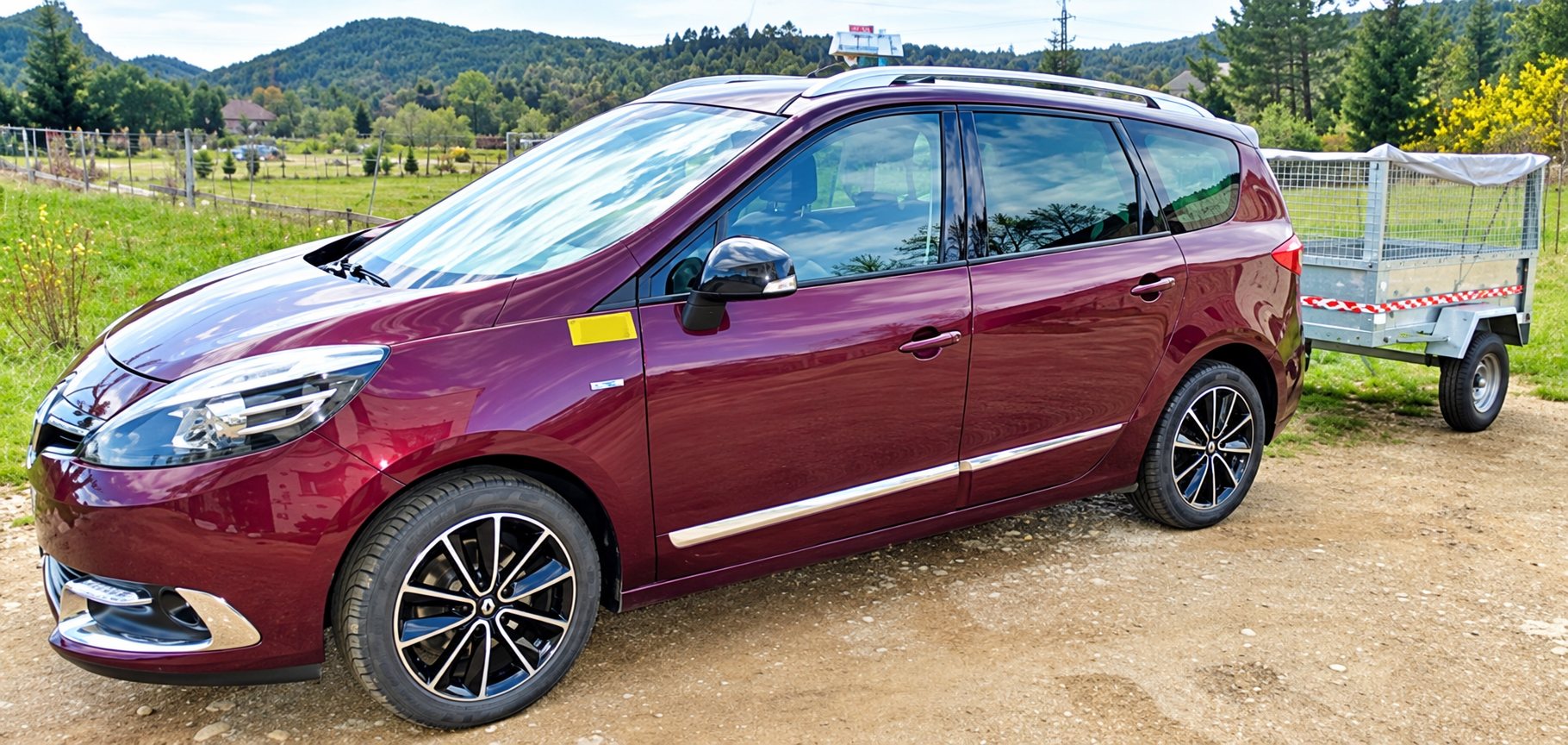 Un monospace bordeaux équipé d'un lumineux de taxi sur le toit est garé sur un chemin de terre et attelé d'une remorque grillagée, avec des collines en arrière-plan.